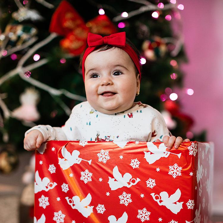 Smiling baby wearing a red headband with a bow, sitting inside a festive red gift box with white holiday-themed designs, in front of a decorated Christmas tree with lights.