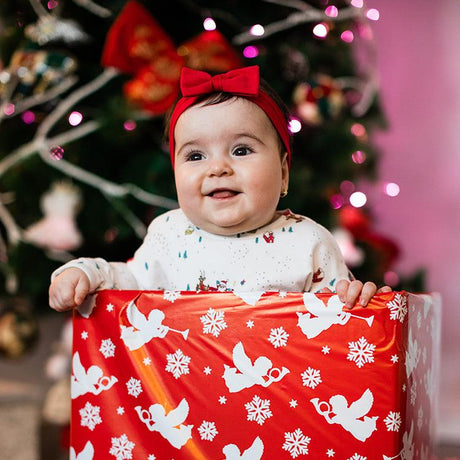 Smiling baby wearing a red headband with a bow, sitting inside a festive red gift box with white holiday-themed designs, in front of a decorated Christmas tree with lights.