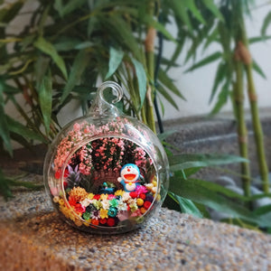Terrarium kaca bulat dengan patung kucing biru di bawah pokok bunga merah jambu di antara bunga awet