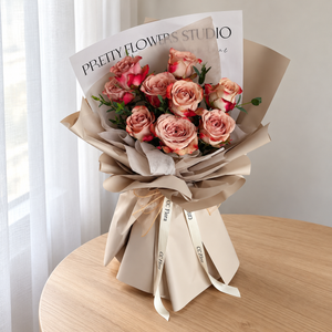 Adore Cappuccino Rose Bouquet in khaki wrapping on a pale oak table beside sheer curtains.
