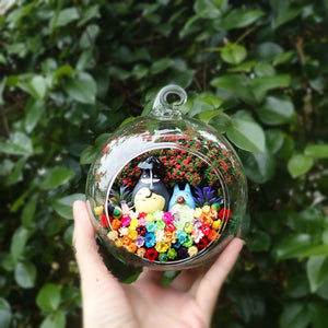Round glass terrarium with grey and blue figurines among preserved flowers