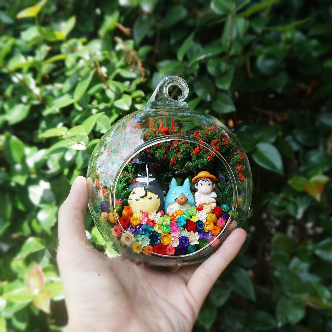 Round glass terrarium with grey and blue figurines with a little girl among preserved flowers