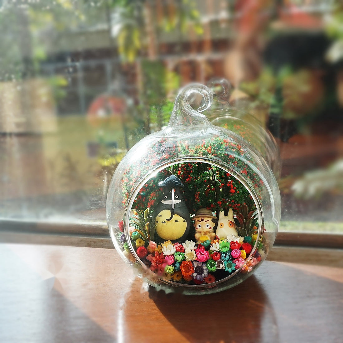 Terrarium kaca bulat dengan patung bulat kelabu dengan hadiah kecil di antara bunga awet