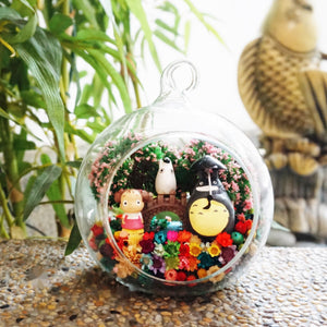 Terrarium kaca bulat dengan patung bulat kelabu bersama rakan kecil di antara bunga awet