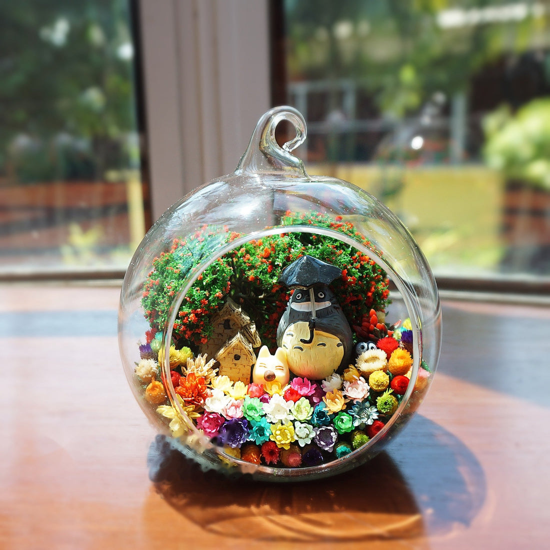 Round glass terrarium with a round grey figurine with an umbrella among preserved flowers