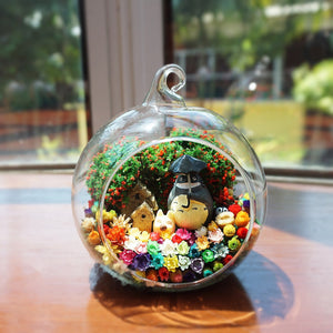 Round glass terrarium with a round grey figurine with an umbrella among preserved flowers