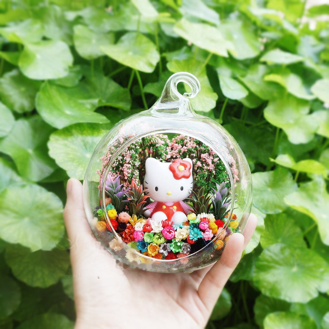 Terrarium kaca bulat dengan patung anak kucing besar bereben di antara bunga awet