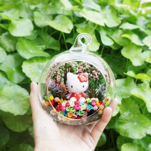 Terrarium kaca bulat dengan patung anak kucing besar bereben di antara bunga awet