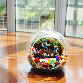 Terrarium kaca bulat dengan patung pasangan tikus berwarna biru di antara bunga awet