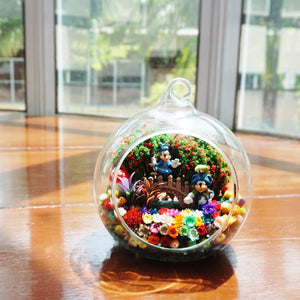 Terrarium kaca bulat dengan patung pasangan tikus berwarna biru di antara bunga awet