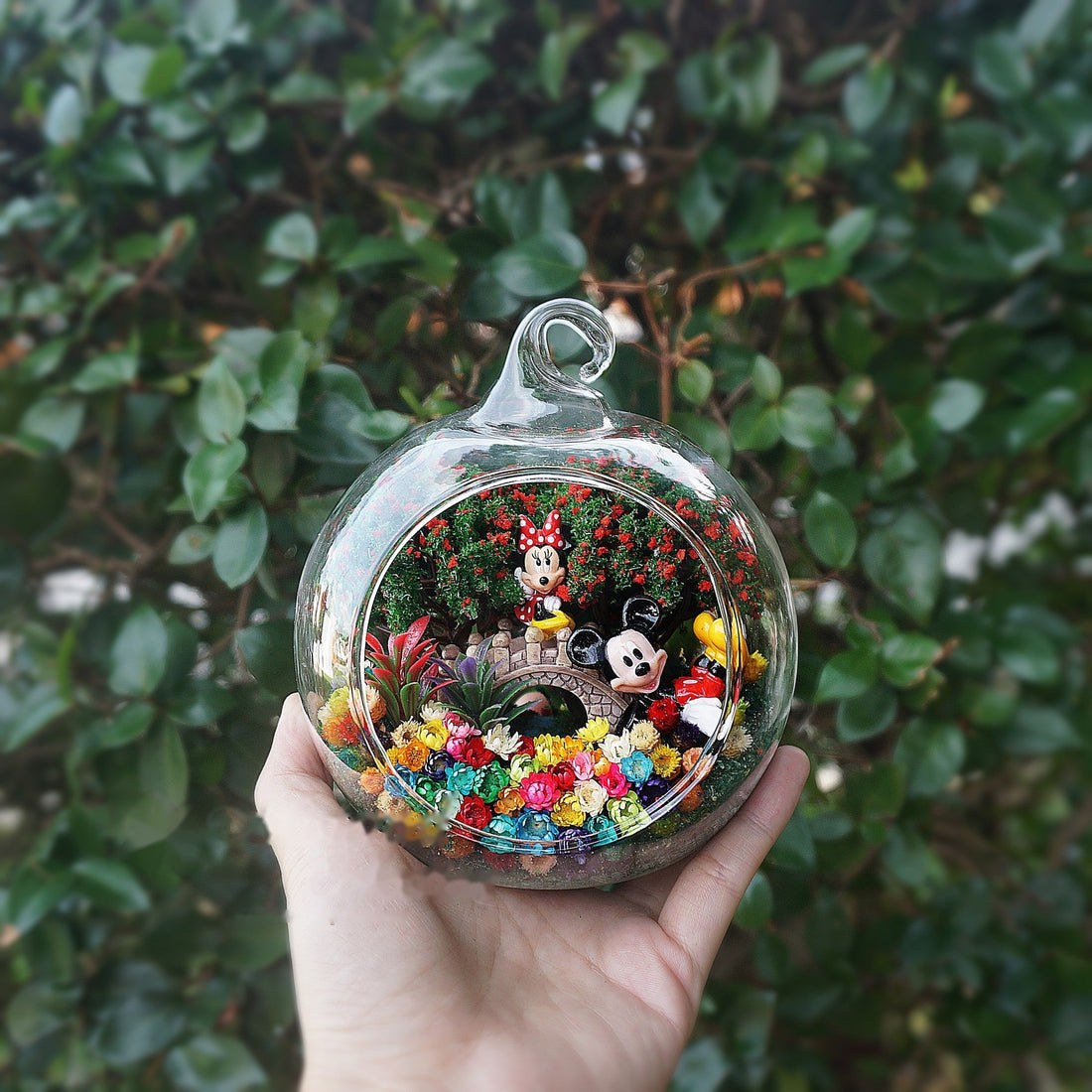 Terrarium kaca bulat dengan patung pasangan tikus berwarna merah di antara bunga awet