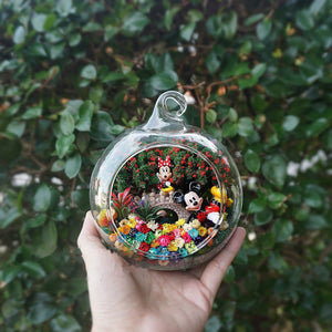 Terrarium kaca bulat dengan patung pasangan tikus berwarna merah di antara bunga awet
