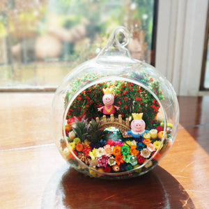 Terrarium kaca bulat dengan patung anak babi merah jambu di antara bunga awet