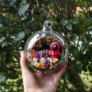 Terrarium kaca bulat dengan patung berpakaian seragam merah jambu di antara bunga awet