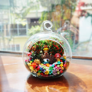 Terrarium kaca bulat dengan dua patung berpakaian overall dan topi di antara bunga awet