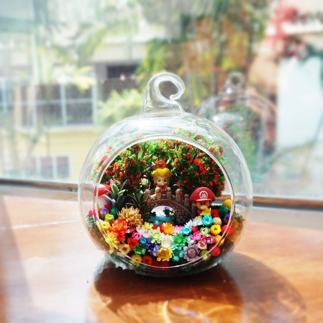 Terrarium kaca bulat dengan dua patung berpakaian overall dan topi di antara bunga awet